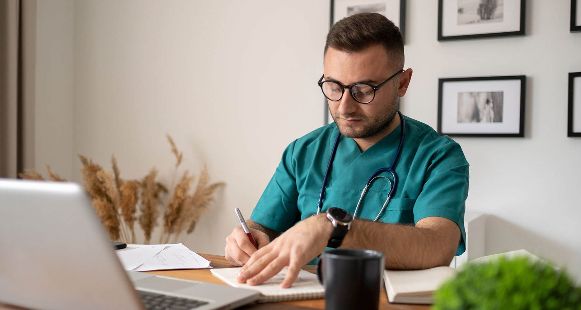 junger, lächelnder Arzt mit Stethoskop sitzt hinter Schreibtisch im Büro