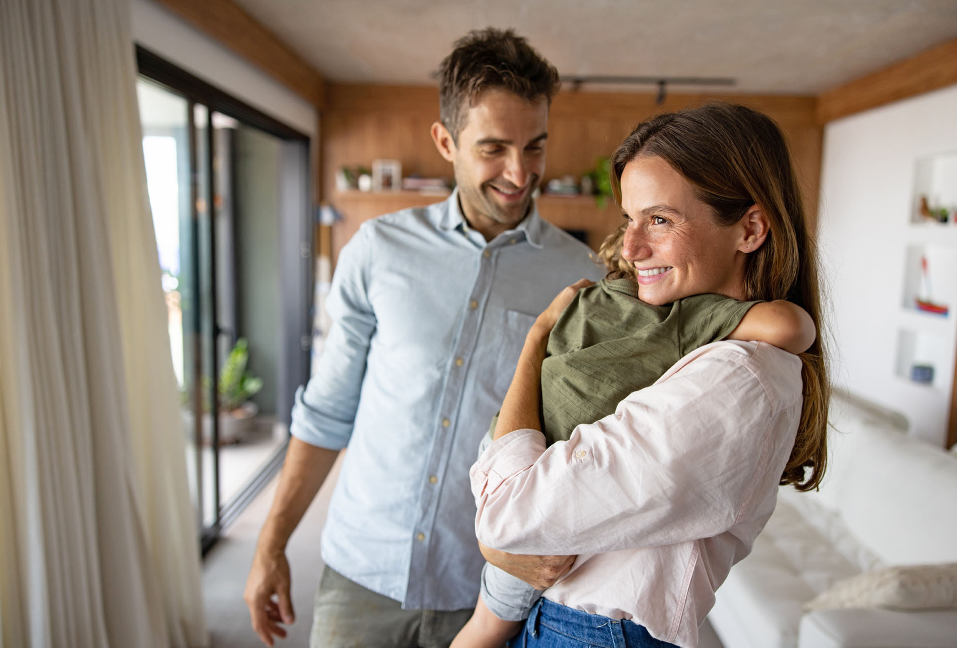 Ein glückliches, junges Ehepaar steht in ihrer Wohnung. Die Mutter umarmt ihr kleines Kind und lächelt.