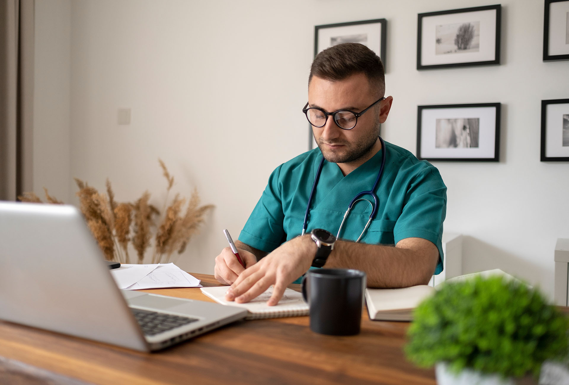 junger, lächelnder Arzt mit Stethoskop sitzt hinter Schreibtisch im Büro